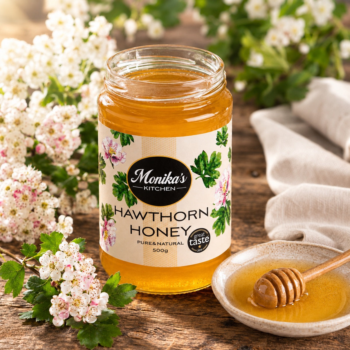 Jar of Monika's Kitchen Hawthorn Honey with a honey dipper on a wooden surface with hawthorn flowers.
