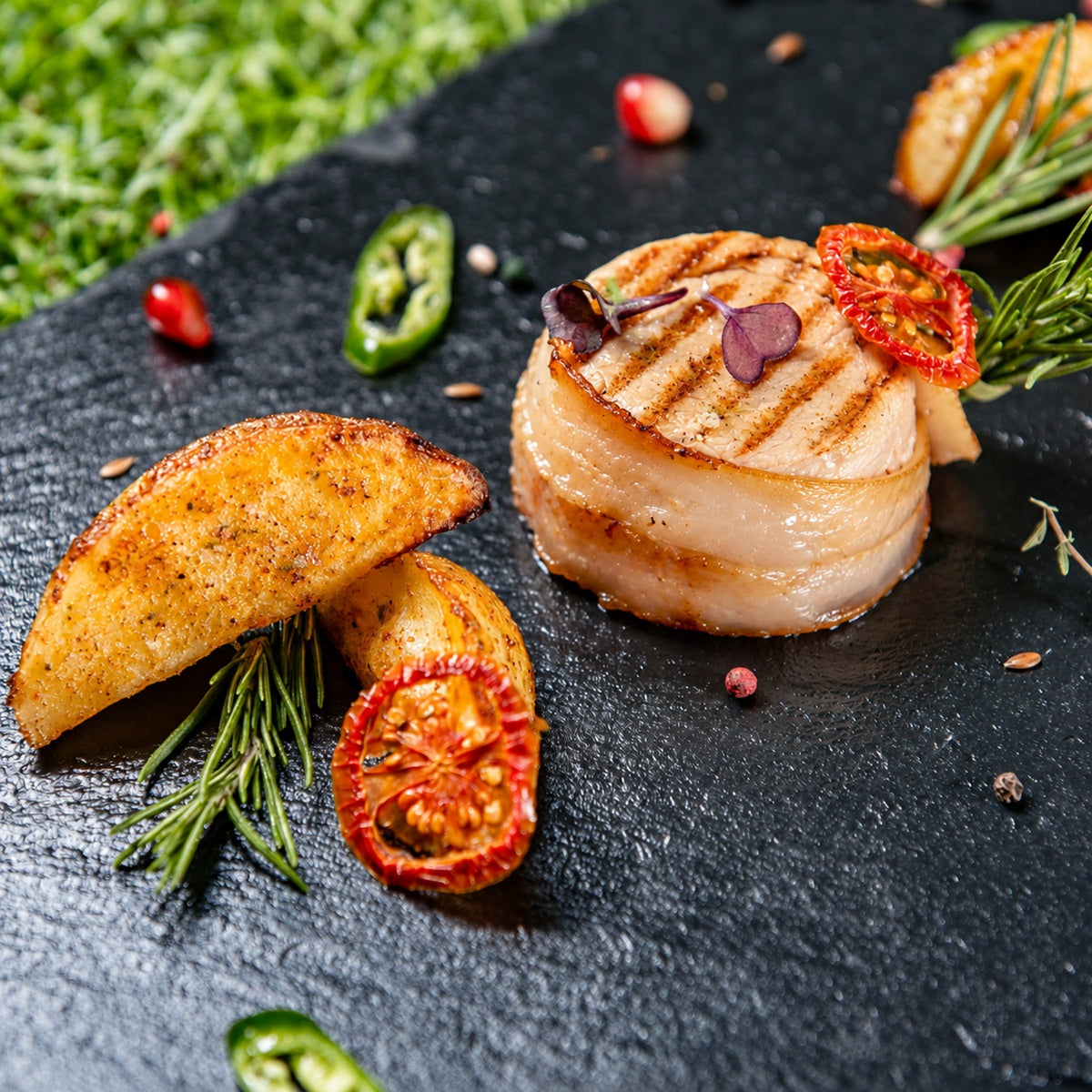 Meat wrapped in smoked Iberico bacon with roast potato wedges on dark slate.