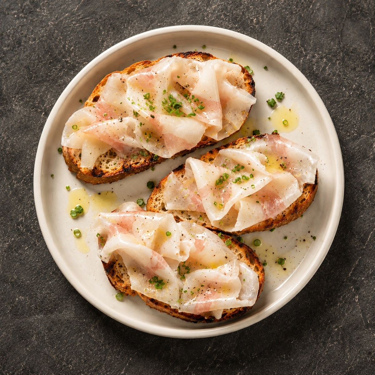 Three slices of toasted bread with slices of lardo and green herbs on a white plate against a dark background.