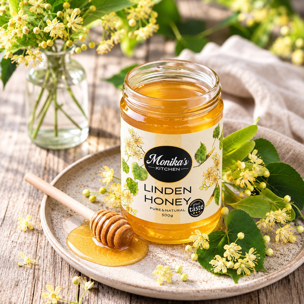 Jar of Monika's Kitchen Linden Honey, also known as Lime Tree Honey on a wooden surface with flowers and a honey dipper.