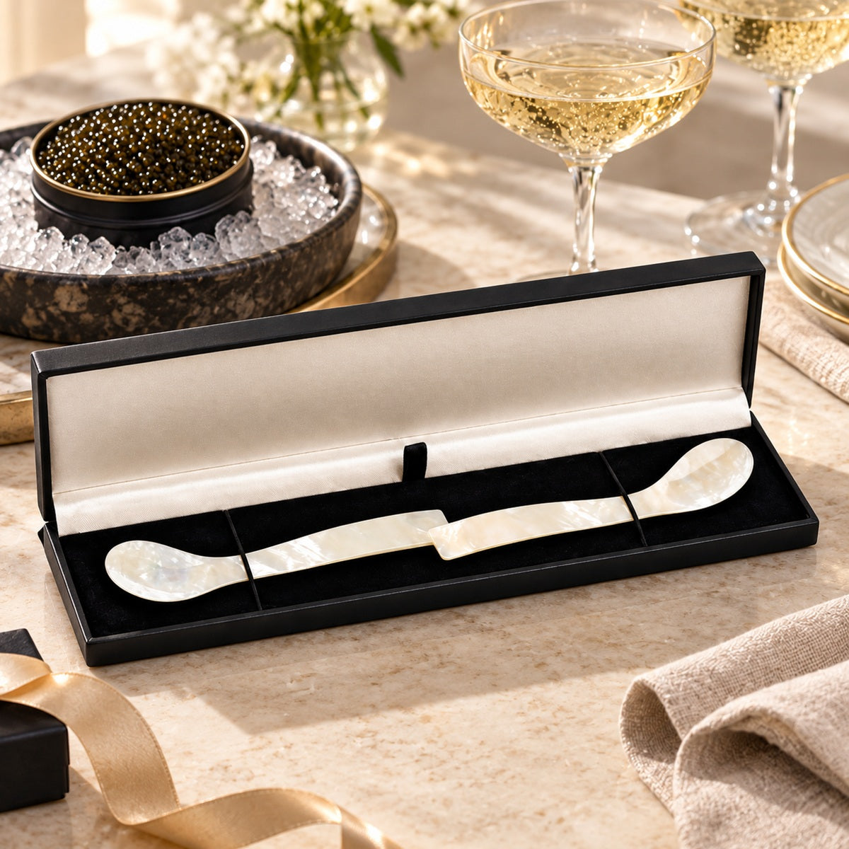 A two-piece mother-of-pearl serving spoon set displayed in an open black gift box with a caviar bowl in the background.