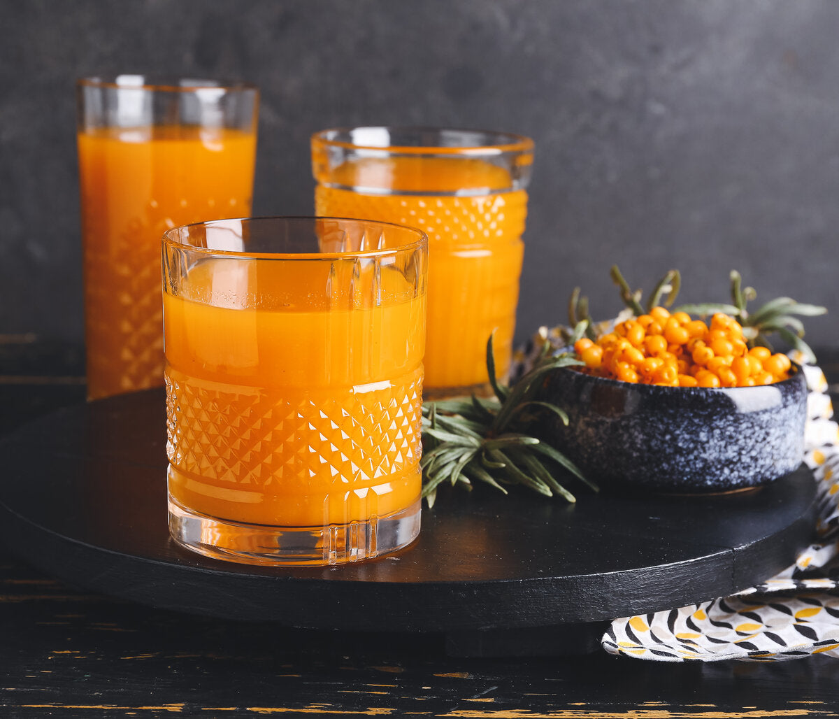 Three glasses of sea buckthorn juice on a dark surface with a bowl of sea buckthorn berries.