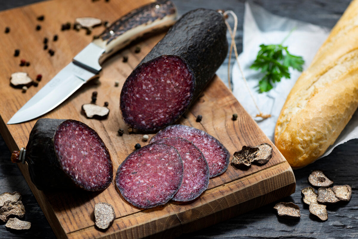 Venison salami with slices of black summer truffles and a fresh baguette on a chopping board.