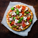 Pizza with bacon, mushroom, tomato, olives and basil toppings on a piece of parchment paper.