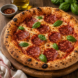 Pizza with beef salami and basil leaves on a wooden board, surrounded by ingredients.