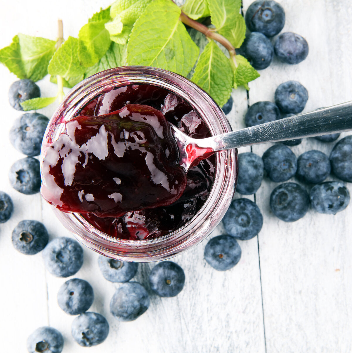 A jar of blueberry jam with a spoon, surrounded by fresh blueberries.