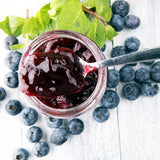 A jar of blueberry jam with a spoon, surrounded by fresh blueberries.