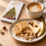 A plate of caramelised white chocolate bars with sliced pear and hazelnuts and a cup of latte with leaf latte art.