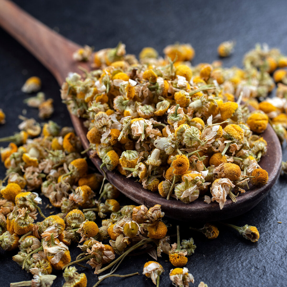 Wooden spoon filled with dried chamomile flowers for use as tea on a dark surface