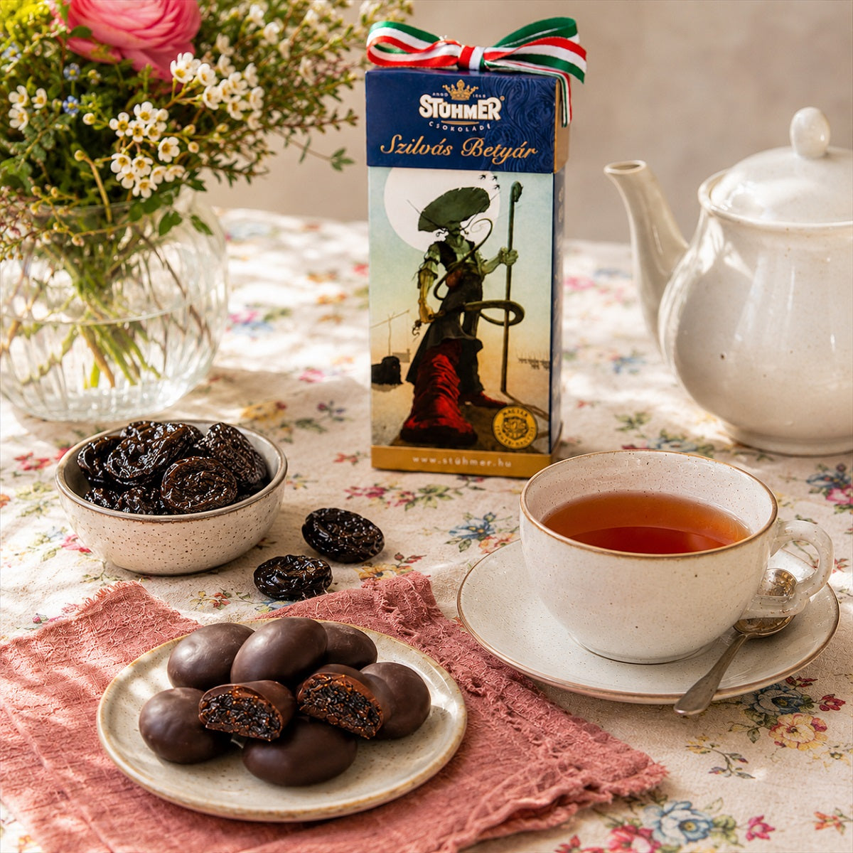 Tea set with a cup of tea, chocolate covered prunes, and a box of Stuhmer Szilvas Betyar on a floral tablecloth.