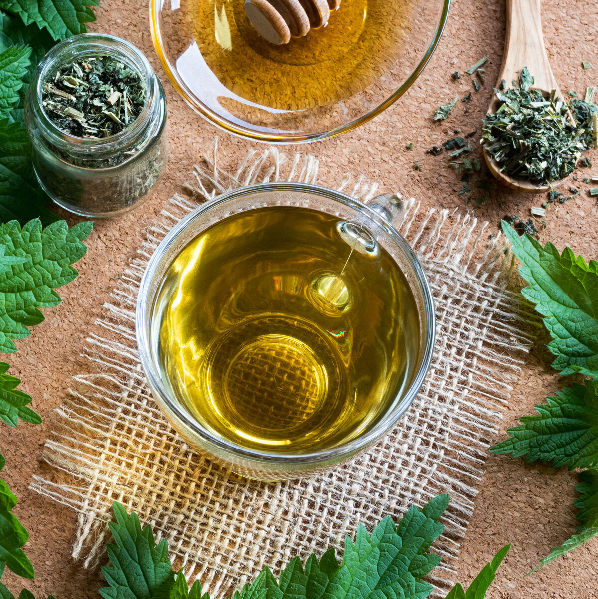 Glass cup of nettle tea, jar of honey, and stinging nettle leaves on a woven mat.