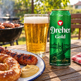 A can of Dreher Gold beer sits on a wooden table beside a glass of golden beer and a plate of grilled sausage.