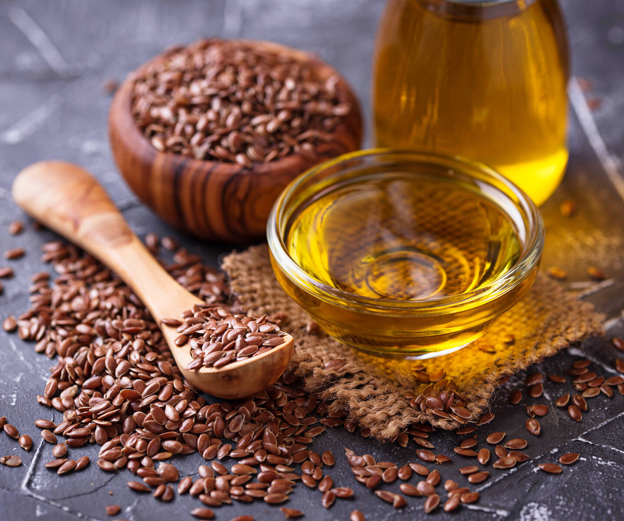 Glass container of flaxseed oil with brown linseeds and wooden spoon on a dark surface