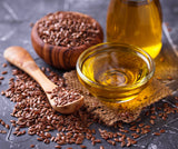Glass container of flaxseed oil with brown linseeds and wooden spoon on a dark surface