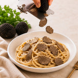 A hand slicing fresh black truffles over a bowl of pasta, with whole black truffles and parsley in the background.