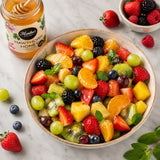 Fruit salad in a bowl with a jar of Monika's Kitchen Hawthorn Honey on a marble surface.