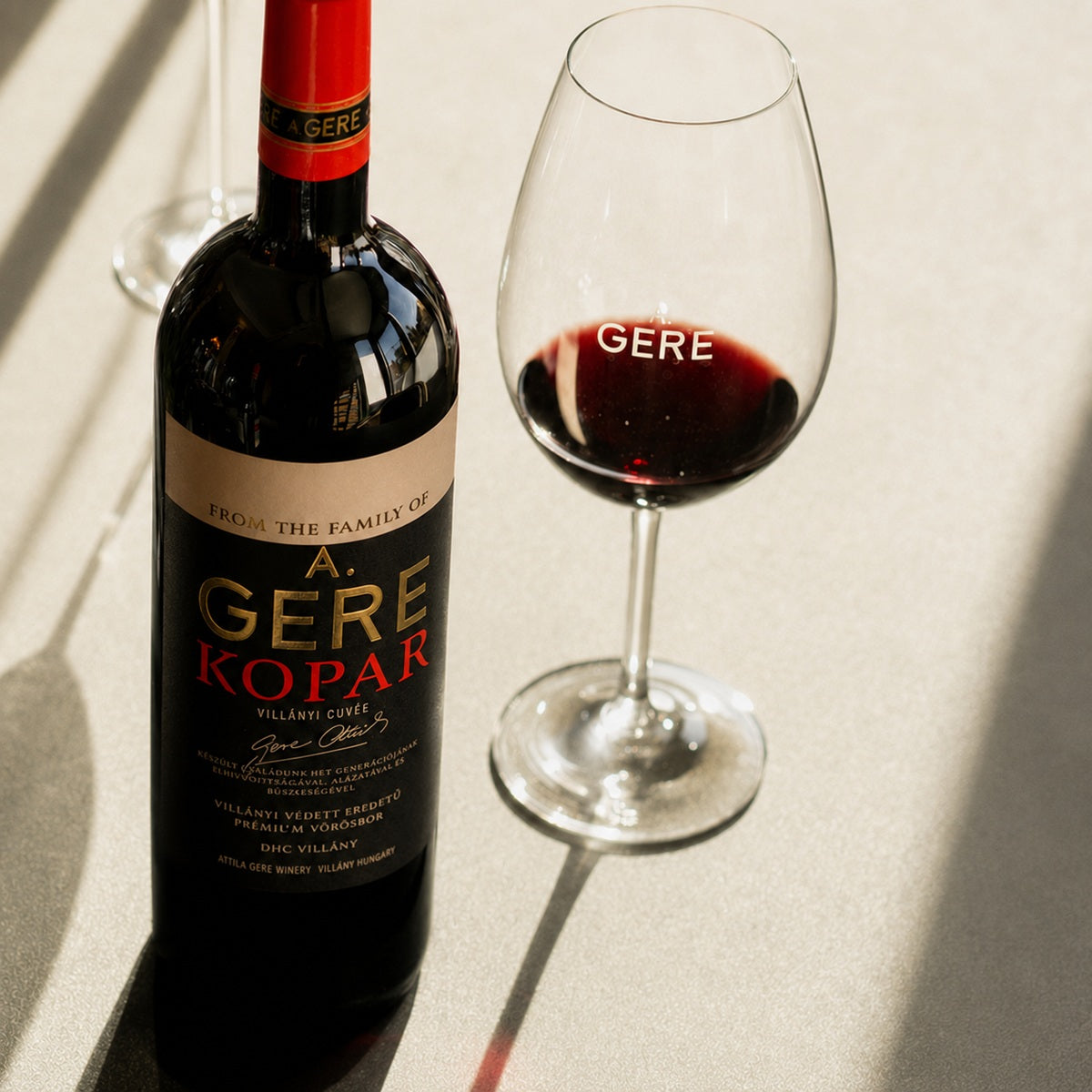 Bottle and glass of A. Gere Kopar Villanyi cuvee red wine on a neutral background.