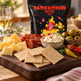 A bag of Flatland Keto Spicy Chilli Crackers on a wooden board surrounded by cheese, salami, olives and a dip bowl.