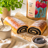 Pastry roll with a ground poppy seed filling on a wooden board with a container of poppy seeds and icing sugar.