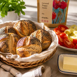 Basket of bread rolls with poppy seeds, a dish of butter, and a plate of sliced tomatoes on a table.