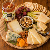 Wooden platter with assorted hard cheeses, fruits, nuts, paired with a jar of chestnut honey on a wooden surface.