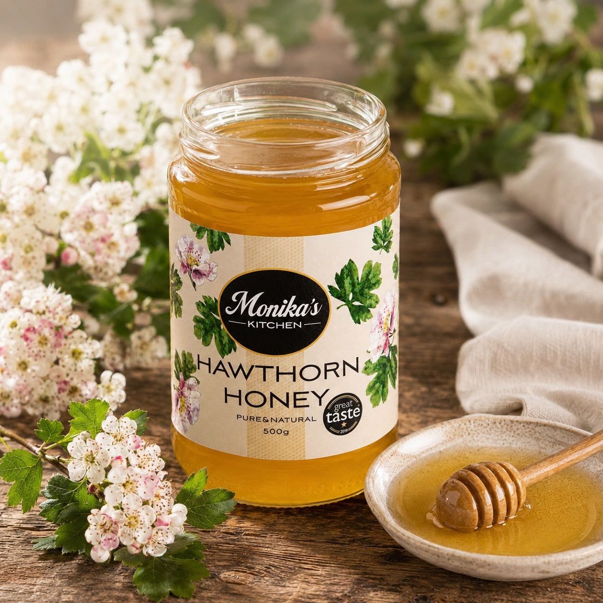 Jar of Monika's Kitchen Hawthorn Honey with a honey dipper on a wooden surface with hawthorn flowers.