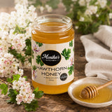Jar of Monika's Kitchen Hawthorn Honey with a honey dipper on a wooden surface with hawthorn flowers.