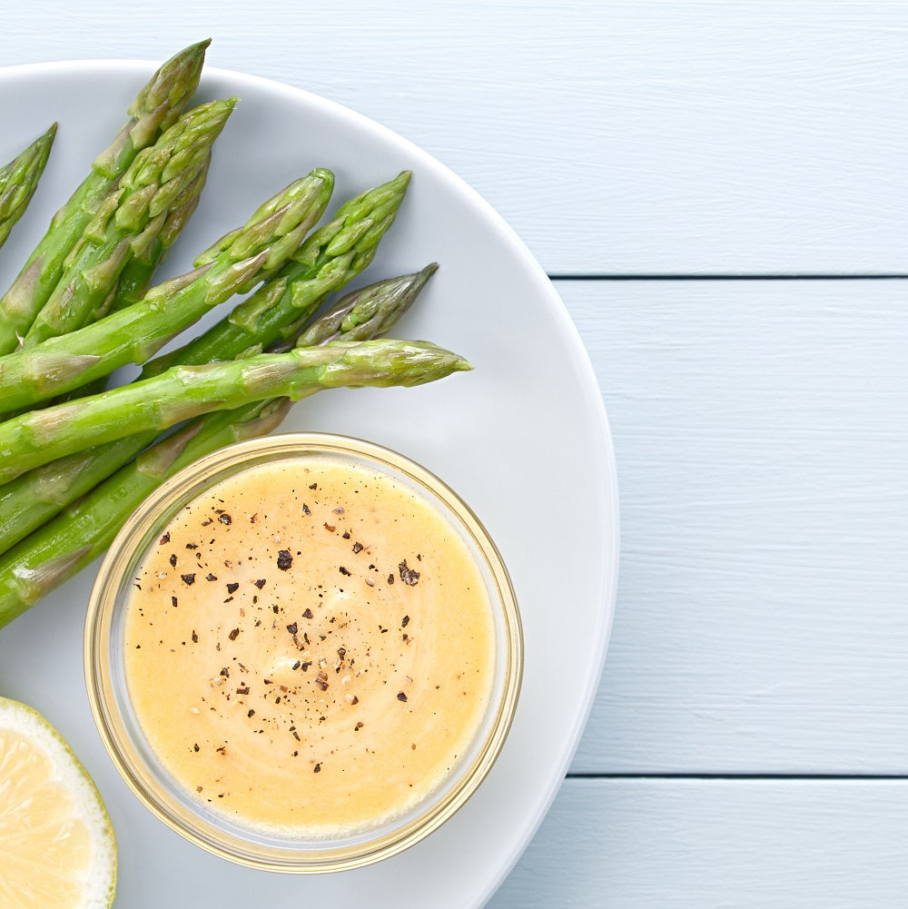 Green asparagus on a white plate with a bowl of Hollandaise sauce sprinkled with ground black pepper.
