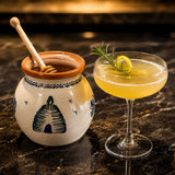 Honey cocktail in a glass with a lemon twist next to honey in a pot on a dark surface.