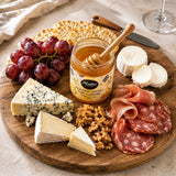 Wooden platter with assorted cheeses, salami, grapes, and a jar of golden honey on a light background.