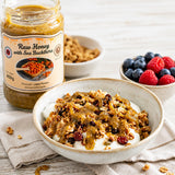 Bowl of granola with yogurt, jar of raw honey with sea buckthorn, and berries on a wooden surface.