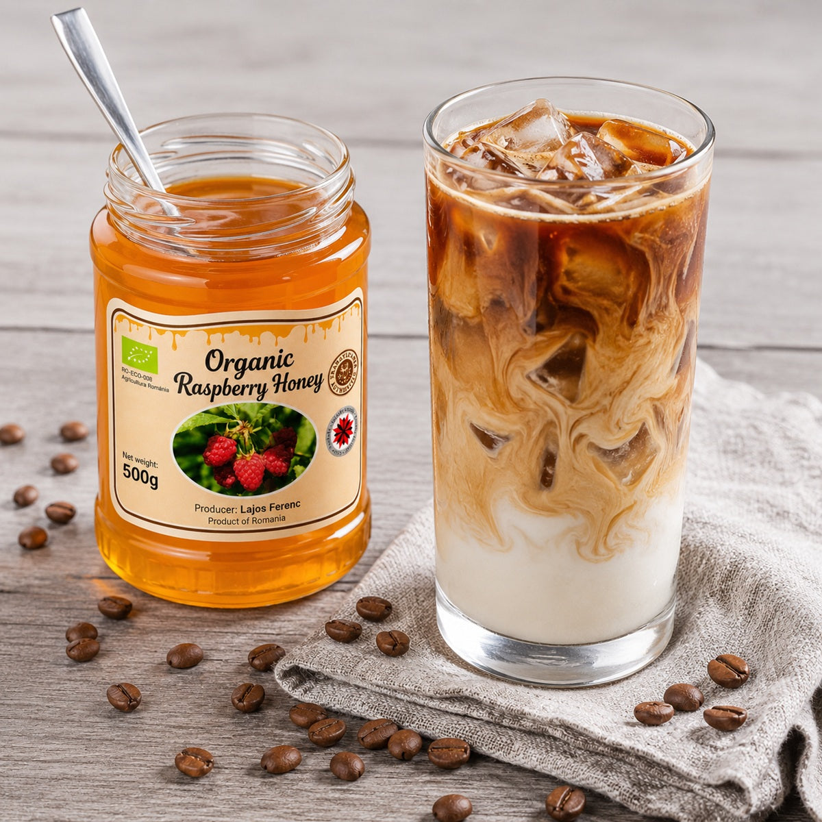 Jar of organic raspberry honey next to a glass of iced coffee with milk on a wooden surface.