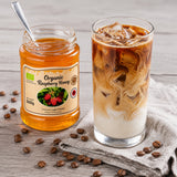 Jar of organic raspberry honey next to a glass of iced coffee with milk on a wooden surface.