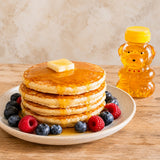 Plate of fluffy, american-style pancakes with honey and berries, with a jar of squeezy honey bear in the background.
