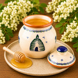 Ceramic honey pot with lid, filled with acacia honey, with a honey dipper, and flowers on a wooden surface