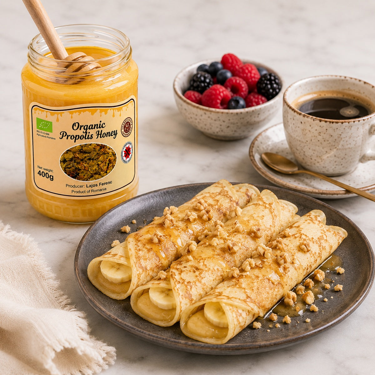 Honey jar with a wooden spoon, crepes on a plate, berries in a bowl, and a cup of coffee on a light surface.