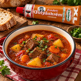 A red enamel bowl filled with a hearty beef and potato goulash, with a tube of Gulyáskrém Csemge in the background.