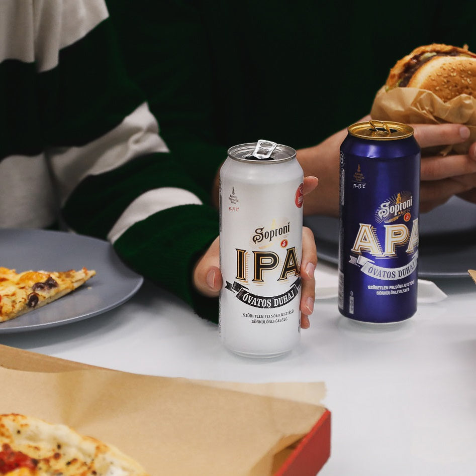 Two cans of Soproni Hungarian beer, one IPA and one APA, held by a person with food on a table.