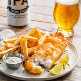 Plated cod in a beer batter with chunky chips, and a glass and can of IPA beer on a wooden table