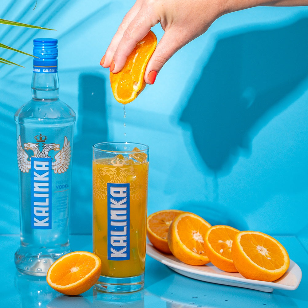 Hand squeezing an orange into a glass of Kalinka vodka with a blue background