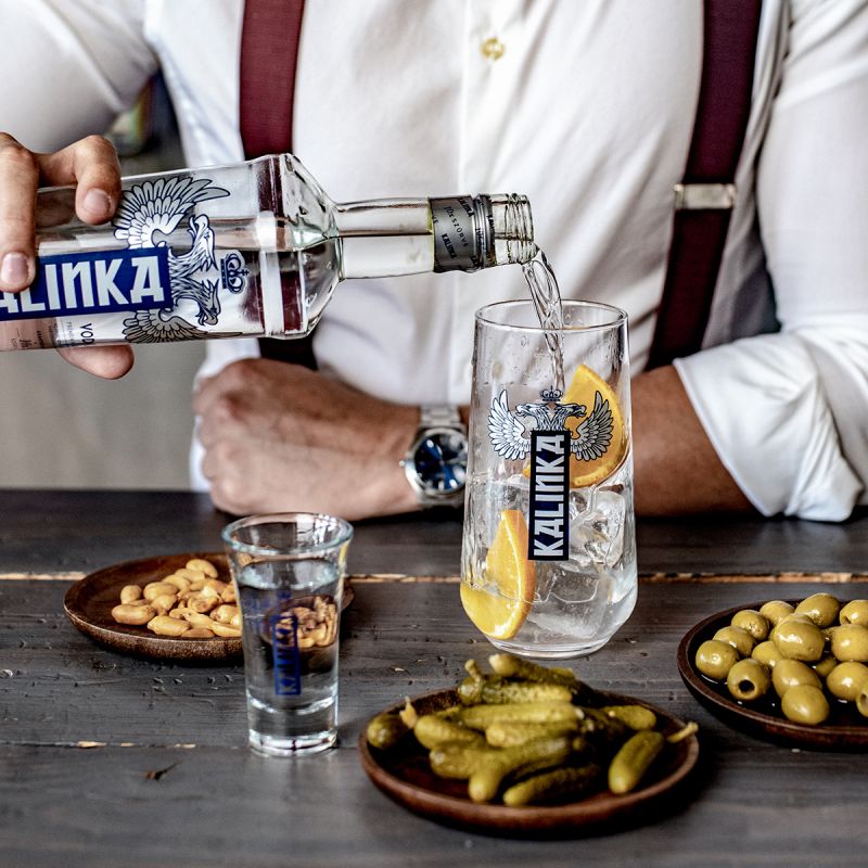 Person pouring Kalinka vodka into a glass with snacks on a wooden table.