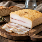 Sliced cured lardo on a wooden board with bread and olives in the background