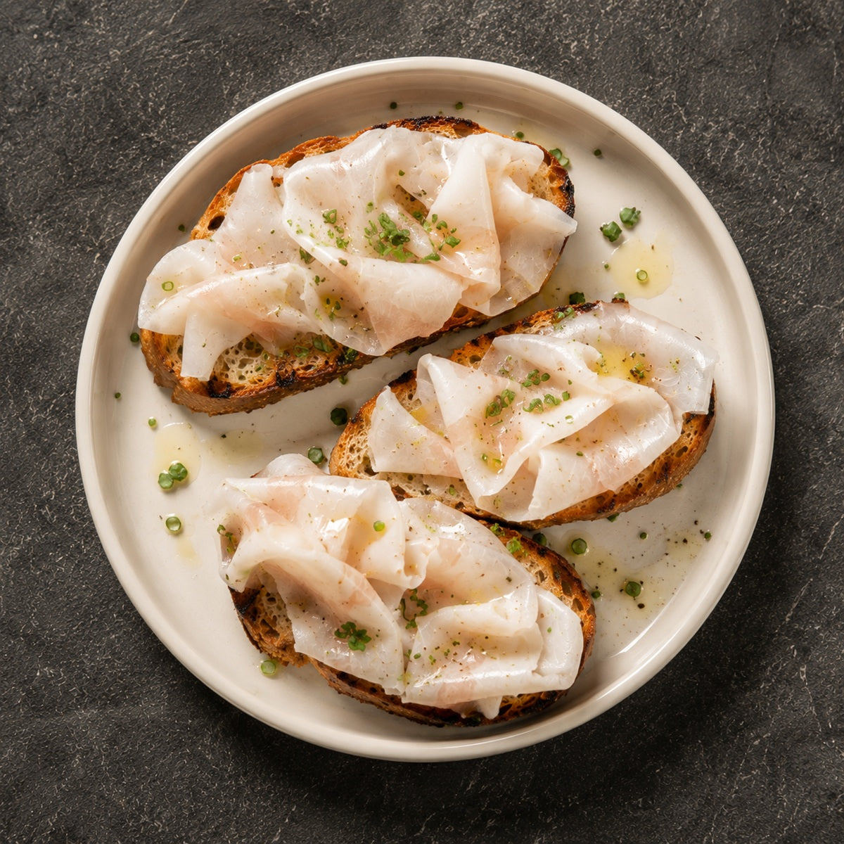 Three slices of toasted bread with slices of lardo and green herbs on a white plate against a dark background.