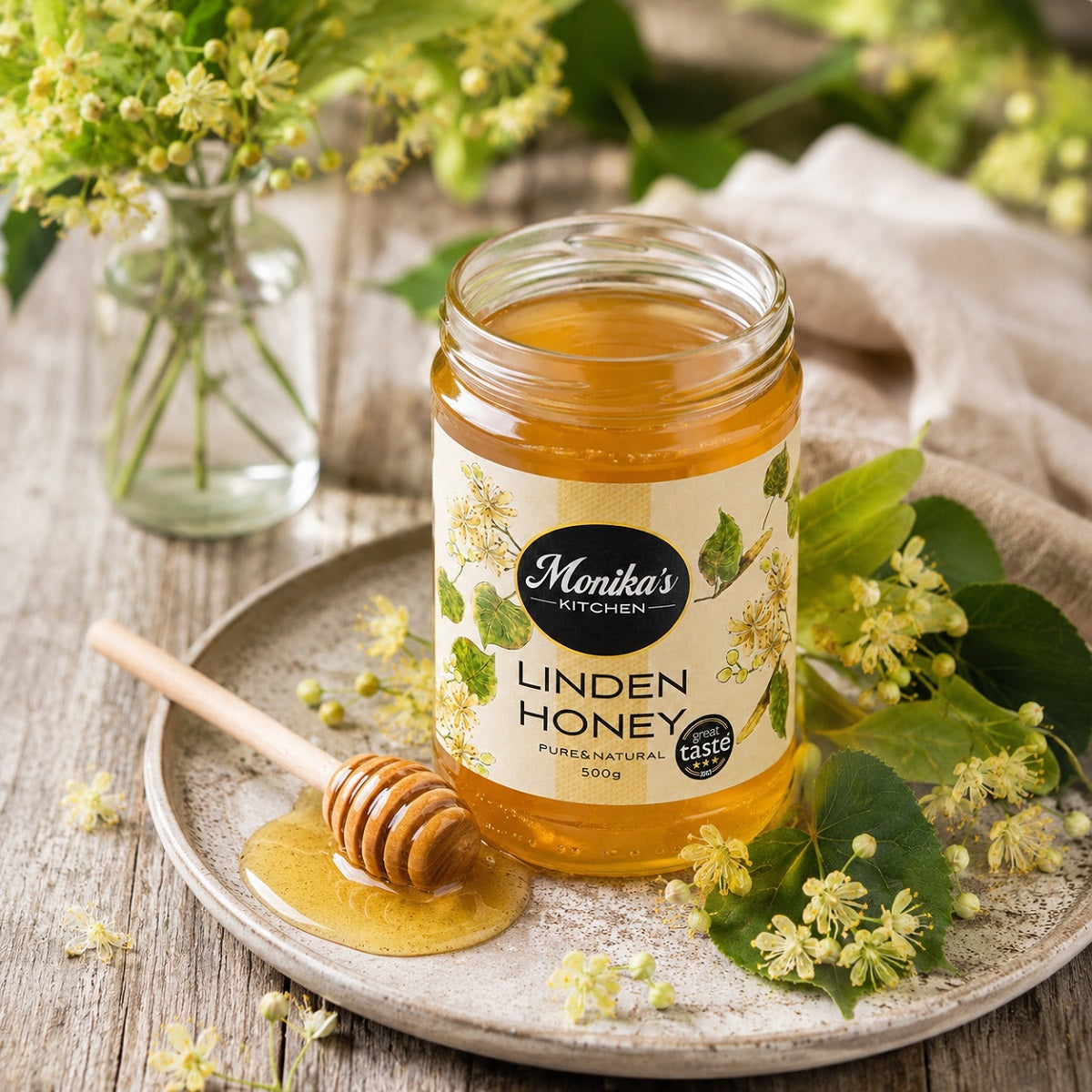 Jar of Monika's Kitchen Linden Honey, also known as Lime Tree Honey on a wooden surface with flowers and a honey dipper.
