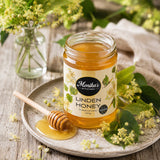 Jar of Monika's Kitchen Linden Honey, also known as Lime Tree Honey on a wooden surface with flowers and a honey dipper.