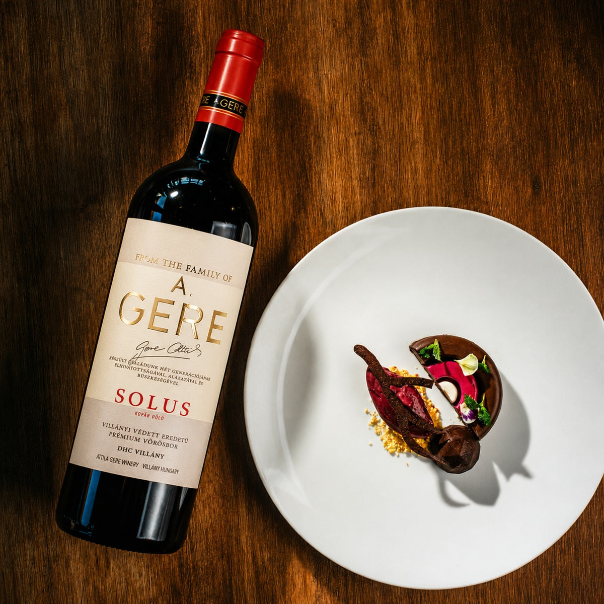 Bottle of A. Gere Solus Merlot wine on a wooden table with a plate of chocolate dessert.