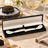 A two-piece mother-of-pearl serving spoon set displayed in an open black gift box with a caviar bowl in the background.