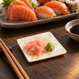 A square white mother-of-pearl dish with green wasabi and pickled ginger and a plate of sushi in the background.