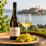 Bottle and glass of oaked chardonnay wine with chardonnay grapes on a wooden oak barrel with a scenic background.
