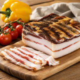 Sliced pancetta bacon from mangalitsa pork on a wooden cutting board with vegetables in the background.